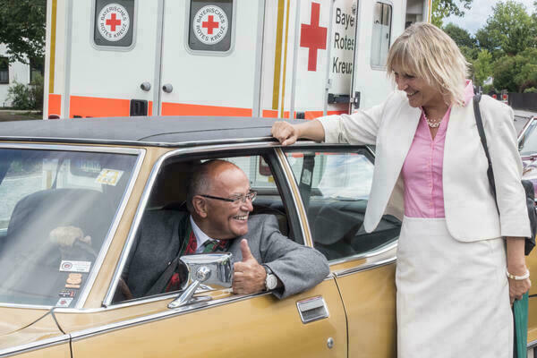Ein Foto vom Bezirkstagspräsidenten Josef Mederer, der in einem senffarbenen Oldtimer sitzt. Neben ihm steht eine blonde Frau. In Hintergrund ist ein Krankenwagen Bayrisches Rotes Kreuz zusehen.
