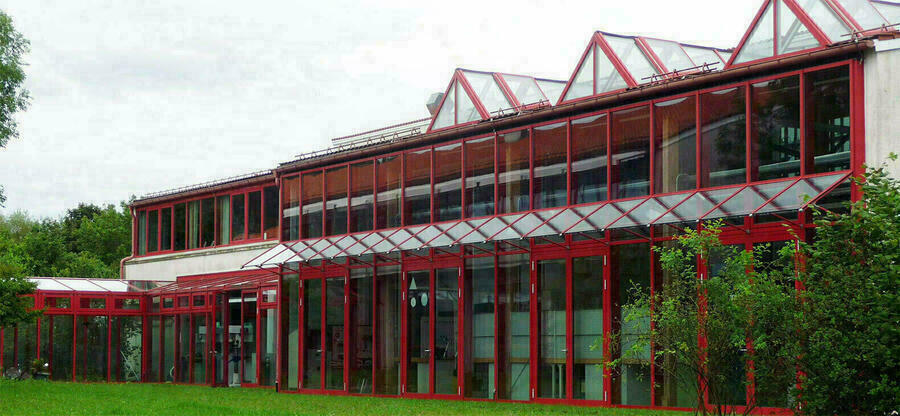 Glasfront der Berufsschule zur sonderpädagogischen Förderung Herzogsägmühle: Ein zweigeschössiger Glasbau mit roten Metallstützen und Glasgauben öffnet sich zu einer Wiese.