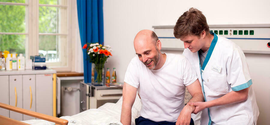 Ein junger Pfleger mit einem weien kurzrmeligen Kittel sttzt den Arm eines Mannes, der auf dem Rand eines Krankenhausbettes sitzt.
