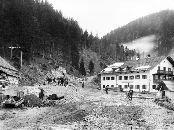 Schwarz-weiß-Foto: Arbeiter schütten neben einem Gasthof eine Kiestrasse auf, die einen Berg hinaufführt. Im Hintergrund sind Wälder und Berge zu sehen.