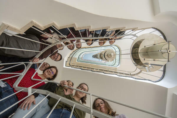 Blick ein Treppenhaus hinauf: Junge Menschen stehen über ein Stockwerk verteilt auf der Treppe und blicken über das Geländer hina.