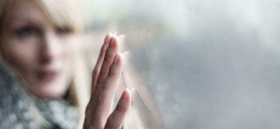 Eine Frau fhrt mit ihren Fingerspitzen ber eine graue Fensterscheibe. Das Gesicht der Frau ist unscharf.