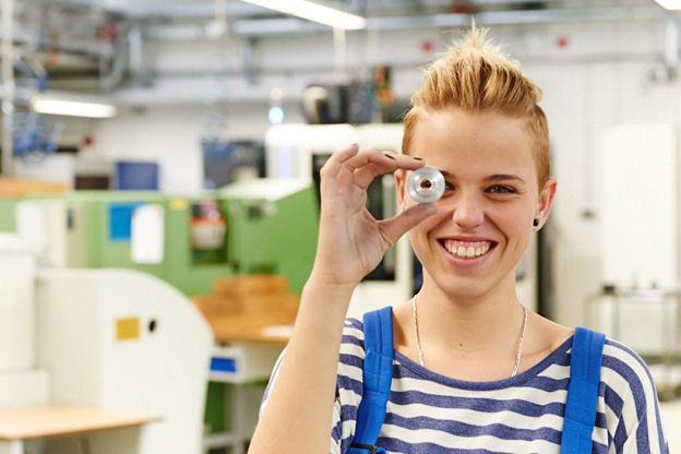Eine lachende junge Frau mit kurzen blonden Haaren und einer blauen Arbeitshose hlt einen Metallring vor das rechte Auge. Im Hintergrund sieht man eine Werkhalle.