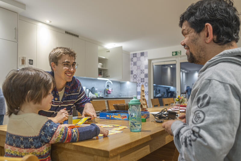 Foto: in einer Küche sitzen zwei Männer un ein Kind an einem Tisch und spielen ein Brettspiel. Die zwei Männer lachen.