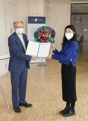 Eine junge Frau mit längeren schwarzen Haaren, blauen Pullover und Maske erhält von einem älteren Mann mit Brille und Maske eine Urkunde und einen Blumenstrauß. 