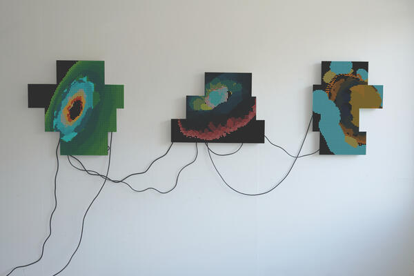 An einer Wand hängen drei Kunstwerke, die durch schwarze Kabel verbunden sind, Sie zeigen mosaikartige abstrakte Formen.