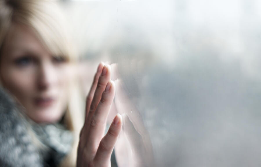 Symbolbild: Eine Frau hat ihre Wange an eine Fensterscheibe gelehnt und streicht mit den Fingern über die Scheibe. Ihre Hand ist im Fokus, sie spiegelt sich in der Scheibe.