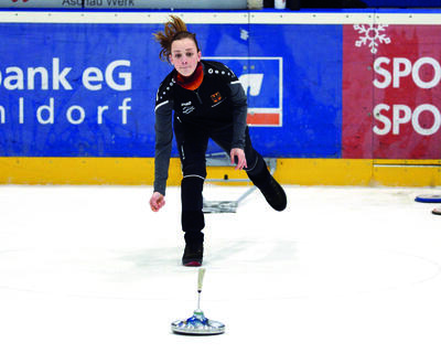 Eine Frau steht auf der Eisfläche in einer Halle und schießt aktiv den Eisstock. Ihr linkes Bein ist dabei nach hinten in der Luft.